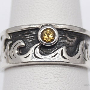 May include: A silver ring with a wave design and a small, round, yellow gemstone. The ring has a textured, dark band with a stylized wave pattern. The gemstone is set in a bezel setting.