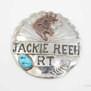 Puede incluir: Un pin redondo de plata y cobre con una piedra turquesa, una figura estilizada y el texto "JACKIE REEH RT".