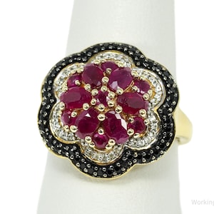 Könnte beinhalten: Goldfarbener Ring mit floralem Design. In der Mitte befinden sich ovale, tiefrote Edelsteine, umgeben von weißen und schwarzen Steinen. Der Ring ist vor einem weißen Hintergrund platziert.