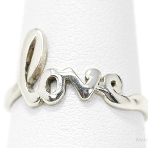 May include: A silver ring spelling out the word "love" in a cursive font. The ring is displayed on a white surface, showcasing the polished metal and the elegant design of the jewelry.