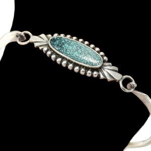 Könnte beinhalten: Ein silbernes Armband mit einem türkisfarbenen Stein als Mittelstück. Der ovale Türkisstein ist von einem perlenbesetzten Rand umgeben und in eine dekorative Silberfassung gesetzt. Das Armband hat ein gebogenes, offenes Design.