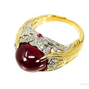 Op de afbeelding: Een gouden ring met een grote, ronde, dieprode edelsteen. De ring heeft een gedetailleerd ontwerp met witte en heldere accentstenen en een gouden band. De ring staat tegen een witte achtergrond.