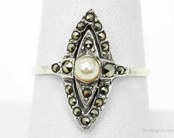 May include: A vintage silver ring featuring a central pearl surrounded by marcasite stones. The ring has an elongated, geometric design with a detailed, ornate setting. The ring is displayed on a white surface.