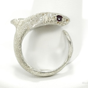 May include: A silver-toned shark ring with a textured finish. The shark's eye is a small, dark red gemstone. The ring is open at the bottom, allowing for size adjustment. The ring is displayed on a white surface.
