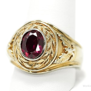 May include: A gold-toned ring featuring an oval, deep red gemstone. The ring has a detailed, textured design around the gem and on the band's shoulders. The ring is displayed on a white background.