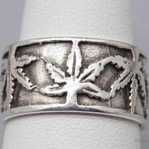 May include: A silver ring featuring a repeating cannabis leaf design. The ring has a wide band with the leaves cut out, creating a textured appearance. The ring is shown on a finger, highlighting the design.