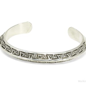 May include: A silver cuff bracelet featuring a repeating geometric pattern of triangles along the outer band. The metal has a polished finish with darkened recessed areas that highlight the engraved triangular motifs. The bracelet is open-ended.