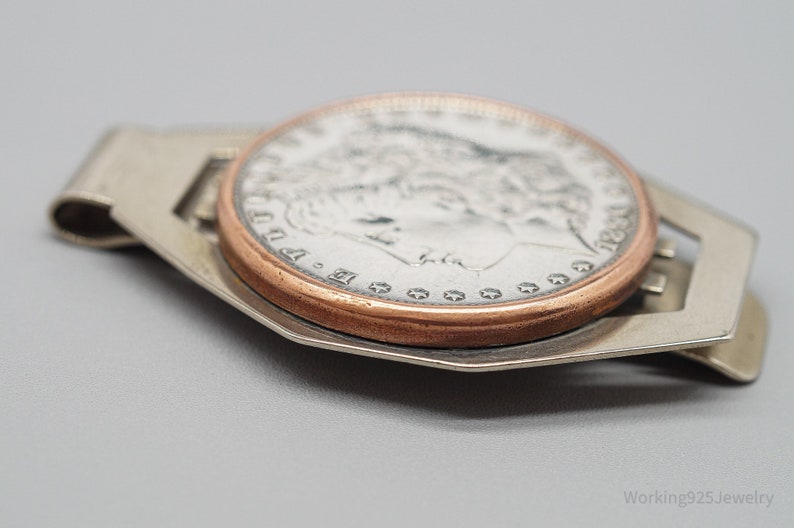 May include: A silver money clip holding a silver coin with a copper-colored rim. The coin features a profile of a woman and the year 1890. The money clip has a rectangular design with cut-out details.