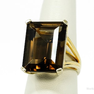 Puede incluir: Un anillo dorado con una gran gema de cuarzo ahumado rectangular. La gema es de un color marrón transparente y rico. La banda del anillo tiene un diseño dividido, agregando un detalle único a la joya.