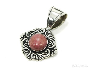 May include: A silver pendant with a square, ornate frame and a round, pink gemstone center. The pendant has a loop for a chain and is set against a white background. The design features swirling patterns.