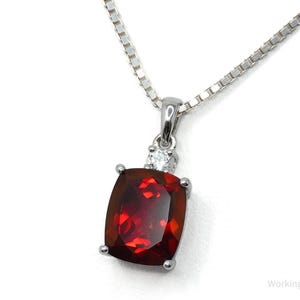 May include: A silver necklace featuring a rectangular, deep red gemstone pendant. The pendant has a smaller, clear stone above the main gem. The chain is a fine, square link style.