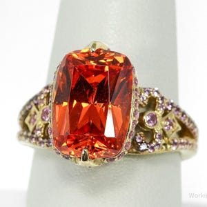 Puede incluir: Un anillo de oro con una gran gema naranja rectangular. El anillo está adornado con gemas rosas más pequeñas y detalles dorados en la banda, creando un diseño lujoso y llamativo.