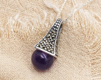 Può includere: Collana con pendente in argento con una gemma sferica viola scuro. Il pendente ha un cappuccio triangolare decorativo in argento con piccoli accenti scuri. La catena della collana è in argento.