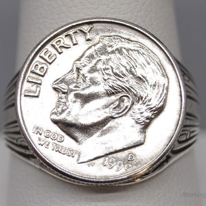 May include: A silver ring with a 1999 dime coin set into it. The coin shows a man's profile, the word "LIBERTY", and the phrase "IN GOD WE TRUST". The ring has a ribbed band design.