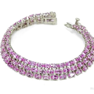Pode incluir: Uma pulseira de prata com três fileiras de pedras rosas. As pedras são retangulares e próximas umas das outras, criando uma faixa contínua de cor. A pulseira está aberta, mostrando o fecho e uma parte da faixa.