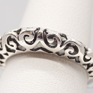 May include: A close-up of a silver ring with an openwork design. The ring features a repeating pattern of swirling and geometric shapes. The ring is on a finger, showcasing the intricate details of the jewelry.