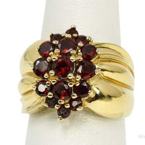 May include: A gold ring featuring a cluster of round, deep red garnet gemstones set in a central arrangement. The band is wide and smooth with a polished finish, tapering slightly toward the back. The stones are held in place by small gold prongs.