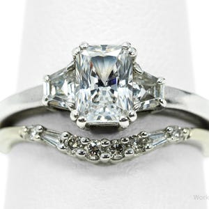 May include: A silver engagement ring set with a radiant cut central stone and two side stones. The accompanying band has a curved design with small, sparkling accents. A classic jewellery set.