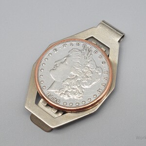 May include: A silver-toned money clip holding a silver coin with a copper-colored rim. The coin features a profile of a woman and the year 1896. The clip has a geometric design and is set against a gray background.