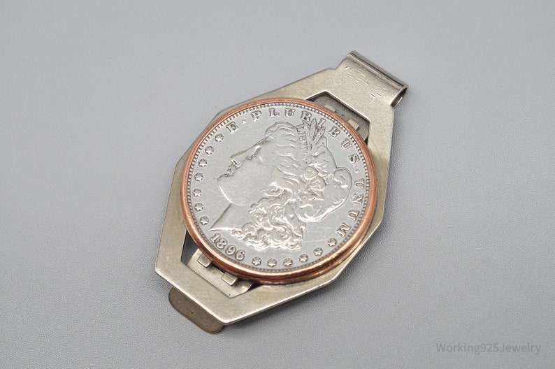 May include: A silver-toned money clip holding a silver coin with a copper-colored rim. The coin features a profile of a woman and the year 1896. The clip has a geometric design and is set against a gray background.