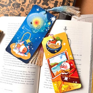 May include: Two rectangular bookmarks with tassels. The blue bookmark features a glowing fish and a sleeping fish on a grid background. The yellow bookmark displays a bowl of ramen and a character sitting in a red chair looking at the ocean.