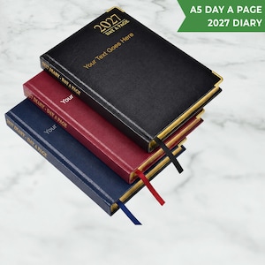 May include: A stack of three A5 day-a-page 2027 diaries in black, burgundy, and navy blue. Each diary features gold-coloured corner accents, gold-edged pages, a ribbon bookmark, and the text 2027 DAY A PAGE and Your Text Goes Here on the cover.