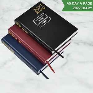 May include: Three A5 day-a-page 2027 diaries stacked vertically in black, burgundy, and navy blue. The top black diary features gold text reading 2027 Day A Page and a white rectangular box with the text PERSONALISED WITH YOUR IMAGE / DESIGN HERE.
