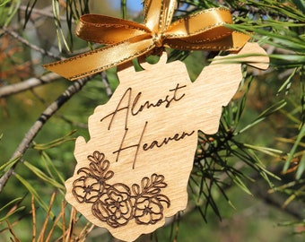 May include: A wooden Christmas ornament shaped like the state of West Virginia with the text "Almost Heaven" and a floral design. The ornament is tied with a gold ribbon.