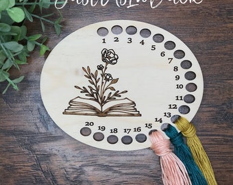 May include: A wooden embroidery thread organizer with 20 holes numbered 1 through 20. The organizer features a design of an open book with a flower growing from it. Three strands of embroidery floss are threaded through the holes.