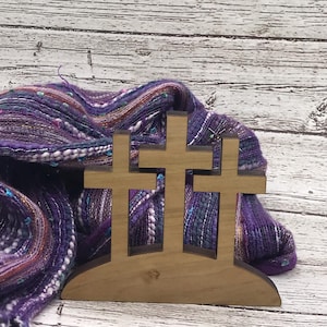 May include: A wooden cross sculpture with three crosses, set against a backdrop of a purple and blue scarf. The crosses are made of light-colored wood. The scarf has a textured appearance.