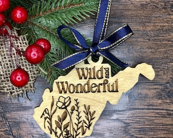 May include: Wooden ornament shaped like the state of West Virginia with the text "Wild and Wonderful" and a floral design. The ornament is decorated with a blue ribbon with gold trim.