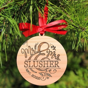 May include: A round wooden ornament with the words "Mr. & Mrs. Slusher Est. 2022" engraved on it. The ornament is decorated with floral designs and is hanging from a red ribbon.