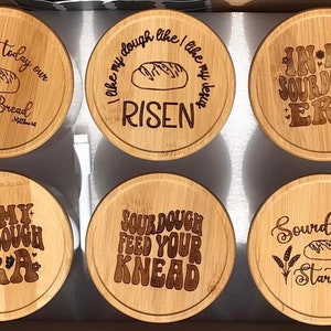 May include: Six round wooden lids with various sourdough-themed sayings engraved on them. The sayings include "Give us today our daily bread", "I like my dough like I like my bread RISEN", "IN-MY SOURDOUGH ERA", "SOURDOUGH FEED YOUR KNEAD", "IN-MY SOURDOUGH ERA", and "Sourdough Starter".