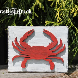 May include: A red wooden crab cutout on a white painted wooden sign. The sign has a distressed white paint finish. The sign reads "Just Us In Duck".
