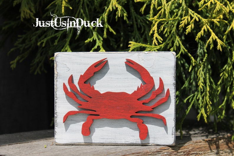 May include: A red wooden crab cutout on a white painted wooden sign. The sign has a distressed white paint finish. The sign reads "Just Us In Duck".