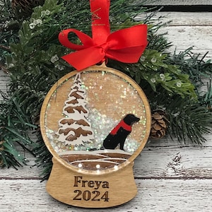 May include: A wooden snow globe Christmas ornament with a black dog silhouette inside. The globe is filled with glitter and a small white tree. The ornament has a red ribbon and the text "Freya 2024" on the bottom.