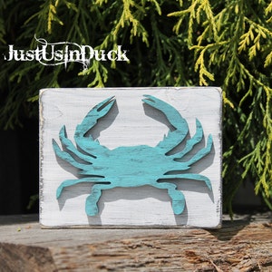 May include: A teal blue wooden crab cutout on a white wooden square sign. The sign has the text "Just Us In Duck" on the top.
