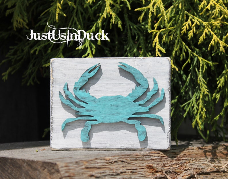 May include: A teal blue wooden crab cutout on a white wooden square sign. The sign has the text "Just Us In Duck" on the top.