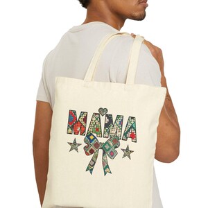 May include: Cream-colored tote bag with the word "MAMA" in colorful patchwork letters. The design includes a heart, stars, and a bow. The bag is carried over the shoulder.