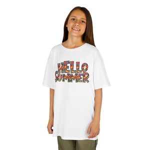 Op de afbeelding: Een wit T-shirt met de tekst "HELLO SUMMER" in kleurrijke, patroonletters. Het shirt wordt door een persoon gedragen, wat een casual, zomerse uitstraling geeft.