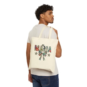 Puede incluir: Un bolso de tela color crema con la palabra MAMA en un diseño de patchwork colorido y variado. El gráfico incluye un pequeño corazón sobre la palabra, un gran lazo decorativo debajo y dos estrellas a cada lado del lazo.