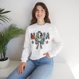 Può includere: Felpa bianca con la scritta "MAMA" in un colorato design patchwork, un cuore, due stelle e un fiocco patchwork. Il top a maniche lunghe è in stile casual.