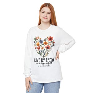 May include: White long-sleeved top with a heart-shaped floral design in red, orange, blue, and yellow. The shirt has the text "LIVE BY FAITH not by sight" and "2 Corinthians 5:7".