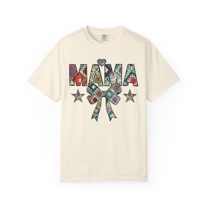 May include: A cream-coloured short-sleeve t-shirt featuring the word MAMA in a colourful patchwork quilt pattern. Above the word is a small heart, and below is a large bow and two stars, all decorated with the same multi-coloured patchwork design.