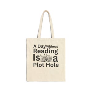 May include: A beige canvas tote bag with two shoulder straps. The front features black text that reads A Day Without Reading Is a Plot Hole, accompanied by a graphic illustration of an open book with flowers growing out of the pages.