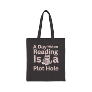 May include: A black tote bag featuring a graphic of a grey cat wearing sunglasses and reading a book. The text printed in light grey reads: A Day Without Reading Is a Plot Hole. The bag has two long shoulder straps and a rectangular body shape.