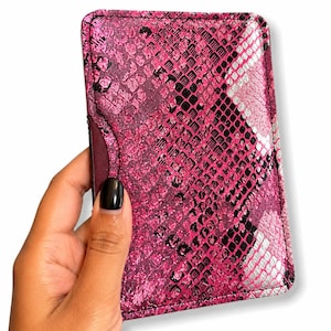 Peut inclure: Un porte-cartes rose avec un motif peau de serpent. Le porte-cartes rectangulaire a des coins arrondis. Le motif peau de serpent est un mélange de rose, de noir et d'argent. Le porte-cartes est tenu dans une main.