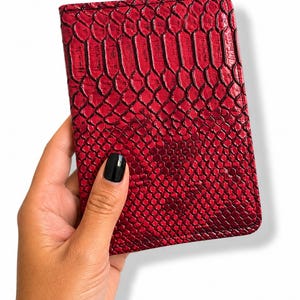 Peut inclure: Un portefeuille rouge avec un motif peau de serpent. Le portefeuille rectangulaire a une surface texturée. Il est tenu dans une main sur fond blanc.