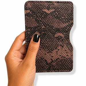 Peut inclure: Un portefeuille marron et noir à motif peau de serpent. De forme rectangulaire avec des coins arrondis et une découpe incurvée sur un côté. La texture gaufrée imite l'aspect réaliste de la peau de serpent, avec des nuances de marron et de noir.