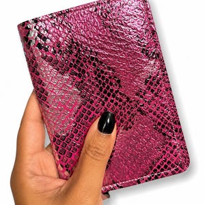 Peut inclure: Un portefeuille rose en faux cuir de serpent avec un motif texturé. Le portefeuille est tenu dans une main avec un ongle peint en noir. Le portefeuille est de forme rectangulaire et a une finition brillante.